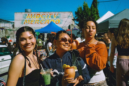 Three women enjoying drinks at a mercado with 'Mid City Mercado' in the background.